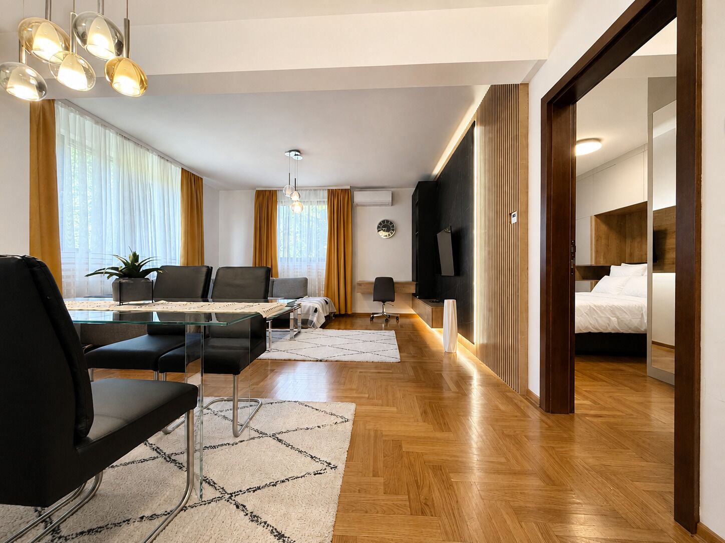 A modern hotel room with a wooden floor, a dining area with a glass table, black chairs, and a large window with curtains.