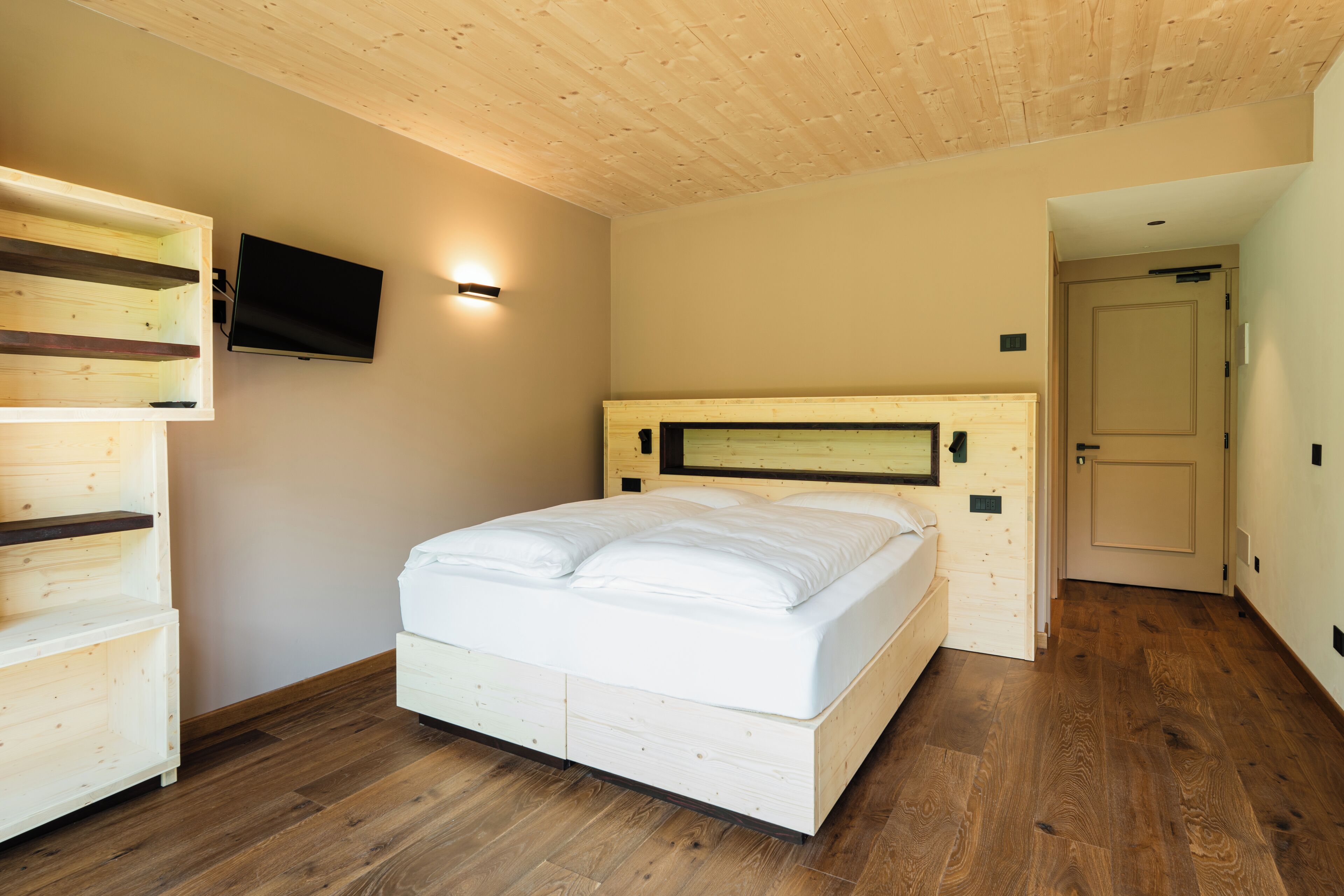 A bedroom with a wooden ceiling, a bed with white bedding, a mounted TV, and a wooden wardrobe.