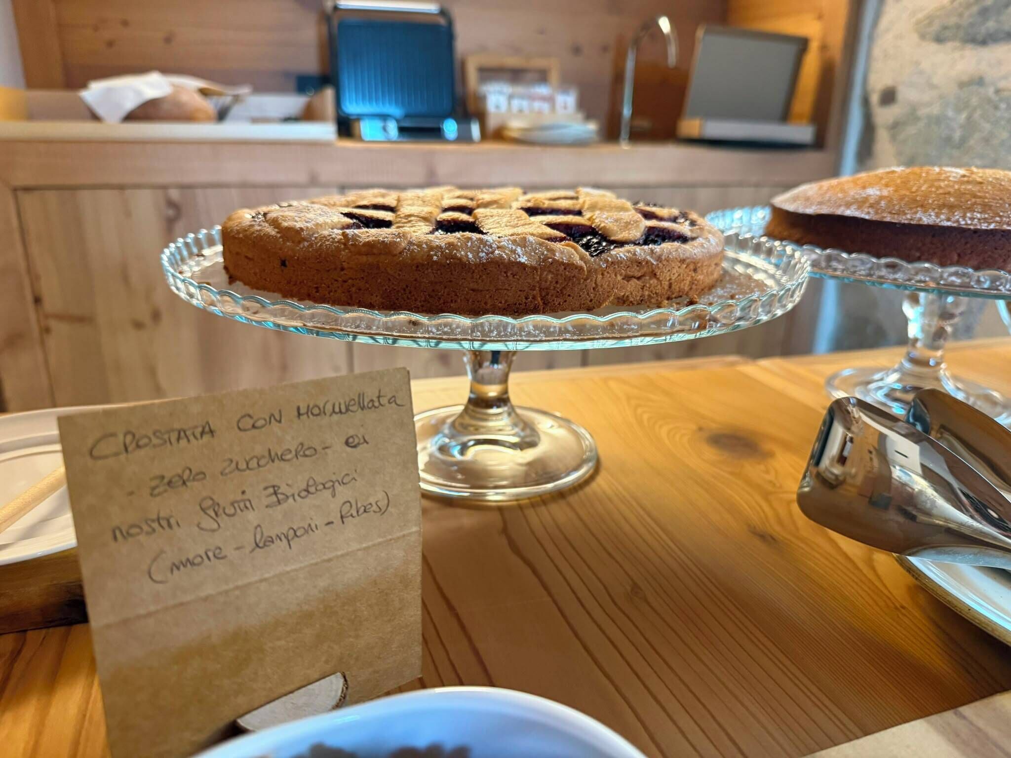 Una vetrina di panetteria con una torta su un piedistallo di vetro, un cartello con scritta a mano e un tavolo di legno.