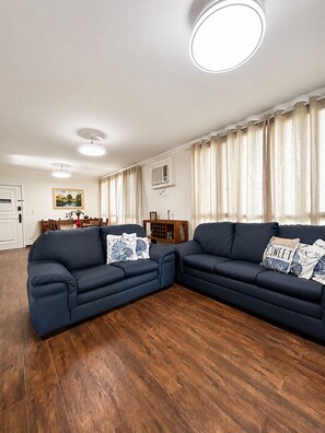 A living room with two dark sofas, a dining table, and a kitchen area in the background. - Cove retreat with 4 bedrooms, pool and comfort. (Guarujá)