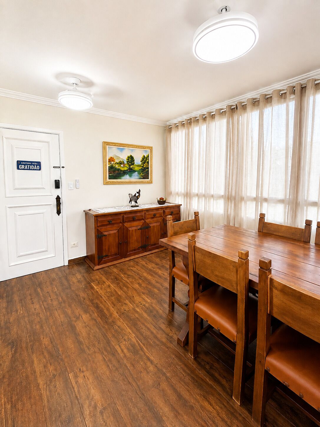 Un comedor con mesa y sillas de madera, una puerta blanca con un letrero, un cuadro en la pared y una ventana con cortinas.