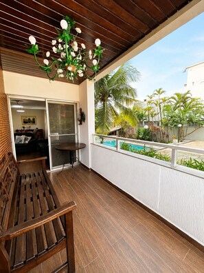 A balcony with a wooden bench, a glass door leading to a room, and a view of a swimming pool and palm trees. - Cove retreat with 4 bedrooms, pool and comfort. (Guarujá)