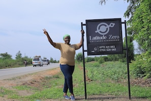 A person standing next to a sign for Latitude Zero Cottages-Kikorongo with contact numbers.