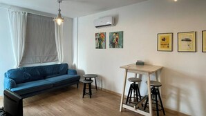 Una sala moderna con un sofá azul, una mesita redonda pequeña y un taburete. Hay cuadros enmarcados en la pared y un equipo de aire acondicionado sobre el sofá.