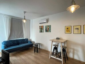 Ein modernes Wohnzimmer mit einem blauen Sofa, einem kleinen runden Tisch und einem Barhocker. An den Wänden befinden sich gerahmte Bilder und über dem Sofa ist eine Klimaanlage angebracht.
