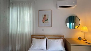 Habitación de hotel con cama, mesita de noche con lámpara y reloj despertador, un cuadro en la pared y un equipo de aire acondicionado.