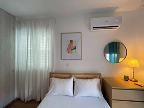 Ein Hotelzimmer mit einem Bett, einem Nachttisch mit Lampe und Wecker, einem gerahmten Bild an der Wand und einer Klimaanlage.