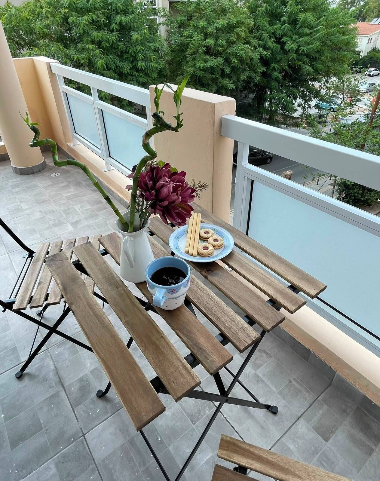 Un balcón con una mesa lista para dos personas, que incluye un jarrón con flores, un plato de galletas y una taza de café.