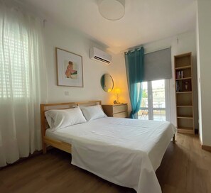 Un dormitorio con cama, mesita de noche, lámpara, estantería y ventana con cortinas.