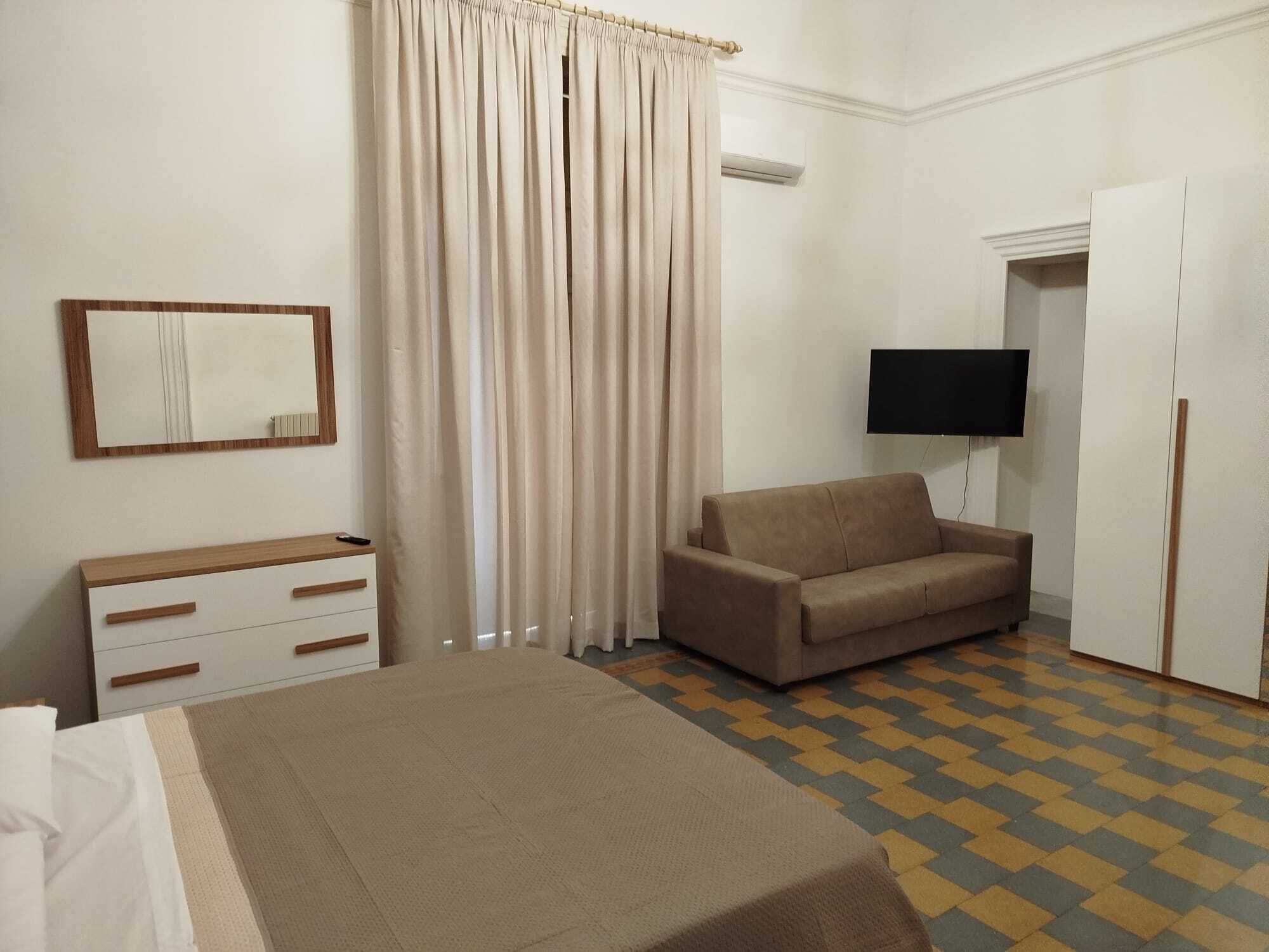 A hotel room with a bed, a sofa, a TV, a mirror, and a window with curtains.