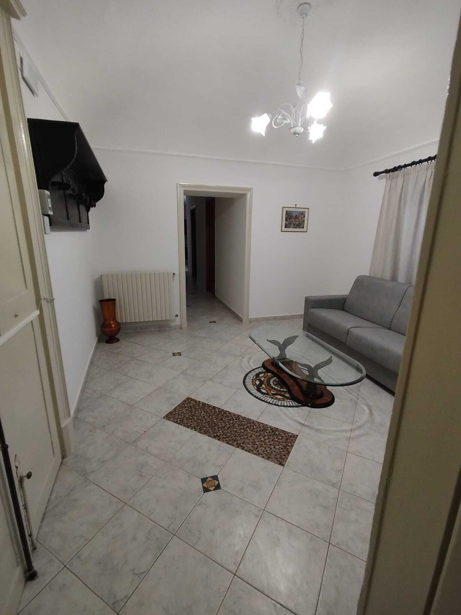 Een woonkamer met een grijze bank, een bijzettafeltje, een radiator en een deur die naar een andere kamer leidt.