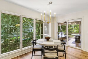 A dining area with a round table, four chairs, a chandelier, and large windows offering a view of greenery outside. - Sunset Rentals | Port Tack 67 (Hilton Head Island)