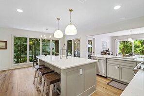 A modern kitchen with a large island, bar stools, pendant lighting, and a view of greenery outside. - Sunset Rentals | Port Tack 67 (Hilton Head Island)