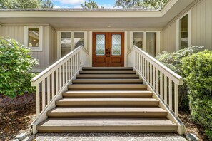 A wooden staircase leading to a double door with glass panels, flanked by white railings and surrounded by greenery. - Sunset Rentals | Port Tack 67 (Hilton Head Island)