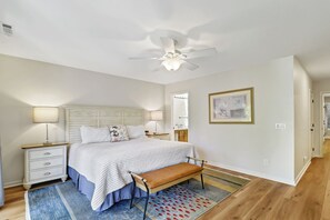 A bedroom with a bed, bedside tables, a bench, a ceiling fan, a bathroom mirror, and a framed picture on the wall. - Sunset Rentals | Port Tack 67 (Hilton Head Island)