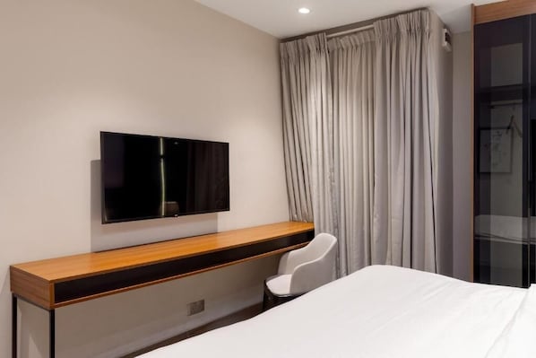 A modern hotel room with a flat-screen TV mounted on the wall, a wooden console table, a single white chair, and a large window with curtains. - Yellow Trumpet Iris Maitama (Abuja)