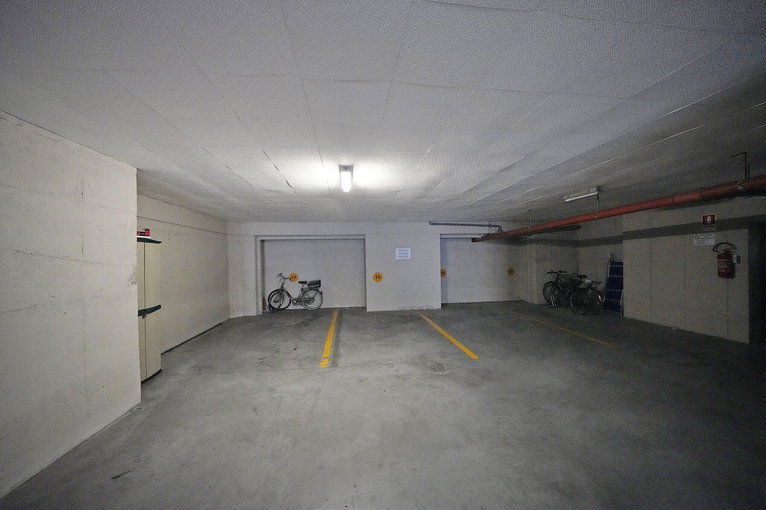 Un parking avec des planchers en béton, des murs blancs et un plafond équipé d’un éclairage fluorescent. On y trouve les portes numérotées 14 et 15, et un vélo est garé près de la porte 14.
