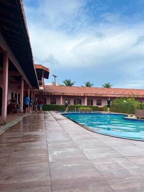A swimming pool area with a covered walkway and a building with arched windows in the background. - Pousada Caranà (Salinópolis)