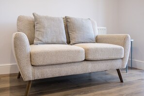 A beige sofa with wooden legs and two pillows, positioned against a wall with a radiator. - Harbourside Haven - stylish marina living (Eastbourne)