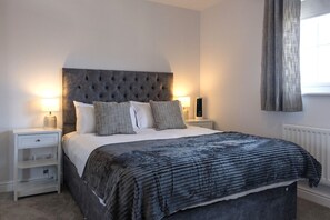 A bedroom with a tufted headboard, a bed with a patterned blanket, two bedside tables with lamps, and a radiator. - Harbourside Haven - stylish marina living (Eastbourne)
