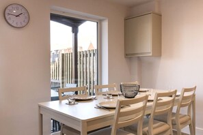 Uma área de jantar com uma mesa posta para quatro pessoas, um relógio na parede e uma janela com vista para edifícios.