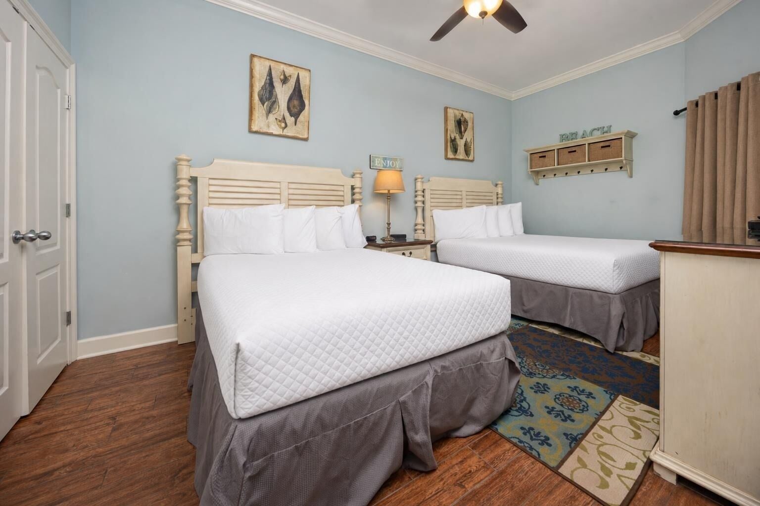 Un dormitorio con dos camas, una mesita de noche, una lámpara, un ventilador de techo y una alfombra decorativa.