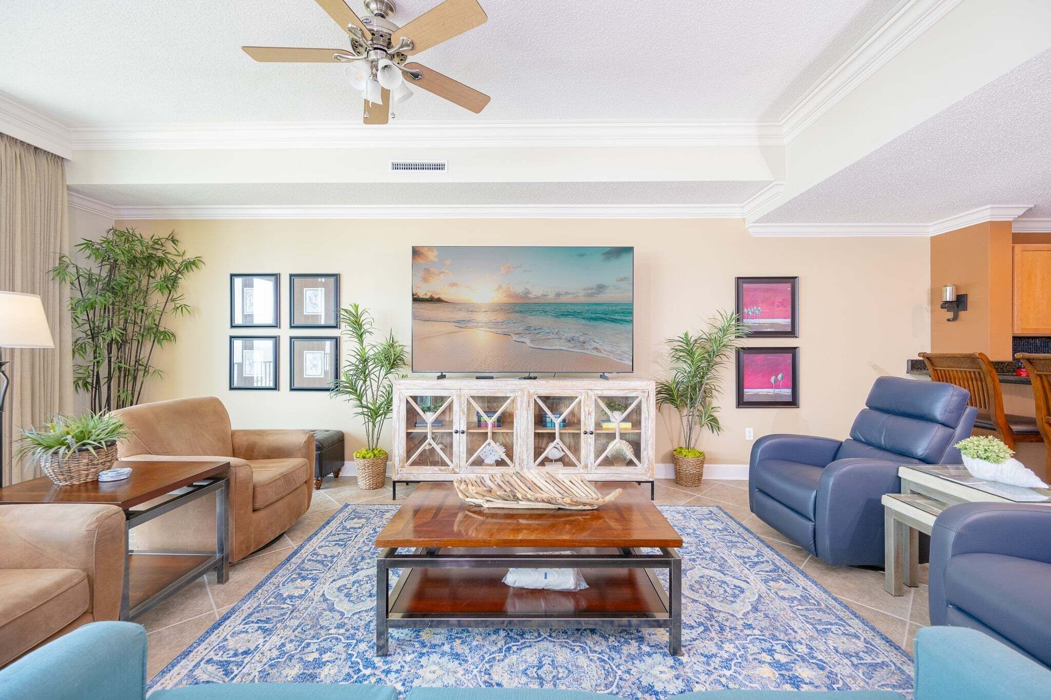 A living room with a ceiling fan, a large painting of a beach, and a dining area with a view of the kitchen.