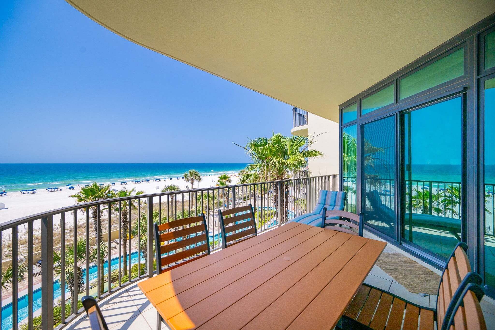 A balcony with a wooden table and chairs, overlooking a beach and a pool.