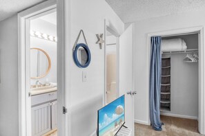 Ein Badezimmer mit Spiegel, einer Meerstern-Dekoration, einem blauen Rundspiegel, einem Fernseher, der eine Strandansicht zeigt, und einem begehbaren Kleiderschrank mit hängender Kleidung.