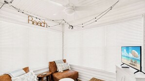 Ein Zimmer mit weißen Jalousien, einem Fernseher, der eine Strandansicht zeigt, zwei Ledersofas mit Kissen und einem Deckenventilator.