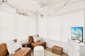 Ein Zimmer mit weißen Jalousien, einem Fernseher, der eine Strandansicht zeigt, zwei Ledersofas mit Kissen und einem Deckenventilator.