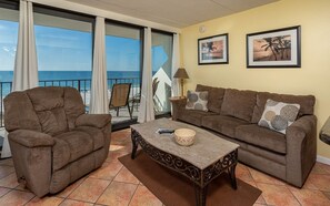 A living room with a sofa, armchair, and coffee table, a balcony with a view of the ocean, and two framed pictures on the wall. - Island Winds East 404 1 Bedroom Condo (Gulf Shores)