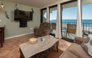 Ein Wohnzimmer mit Fernseher, einem großen Fenster mit Blick auf den Strand, einer Couch und einem Esstisch.