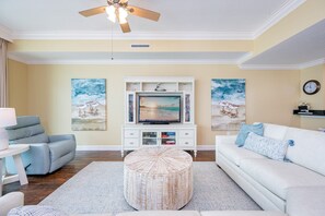 A living room with a flat-screen TV, a ceiling fan, a round ottoman, and artwork on the walls. - Phoenix West 604 3 Bedroom Condo (Orange Beach)