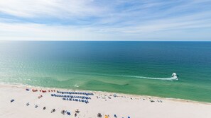 Ein Strand mit klarem türkisfarbenem Wasser, ein sich vom Ufer entfernernde Bootsfahrt und Reihen von Sonnenschirmen und Liegestühlen.