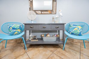 A room with a grey console table, two blue chairs with patterned cushions, a 'WELCOME' sign, a decorative mirror, and a small bird figurine. - Island Winds East 108 2 Bedroom Condo (Gulf Shores)