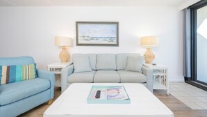 A living room with a light blue sofa, a white coffee table with a magazine, and two side tables with lamps. - Island Winds East 309 2 Bedroom Condo (Gulf Shores)