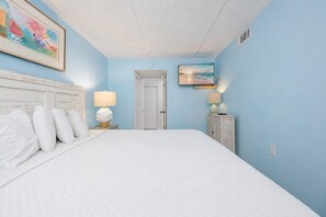 A hotel room with a large bed, a nightstand with a lamp, a painting on the wall, and a television. - Island Winds East 309 2 Bedroom Condo (Gulf Shores)