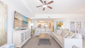 A spacious living room with a large flat-screen TV, a ceiling fan, and a view into the kitchen. - The Oasis 706 Formerly Phoenix West Ii 706 3 Bedroom Condo (Orange Beach)
