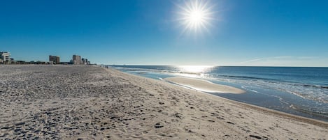Une plage parsemée d’empreintes, un ciel dégagé et des bâtiments en arrière-plan.