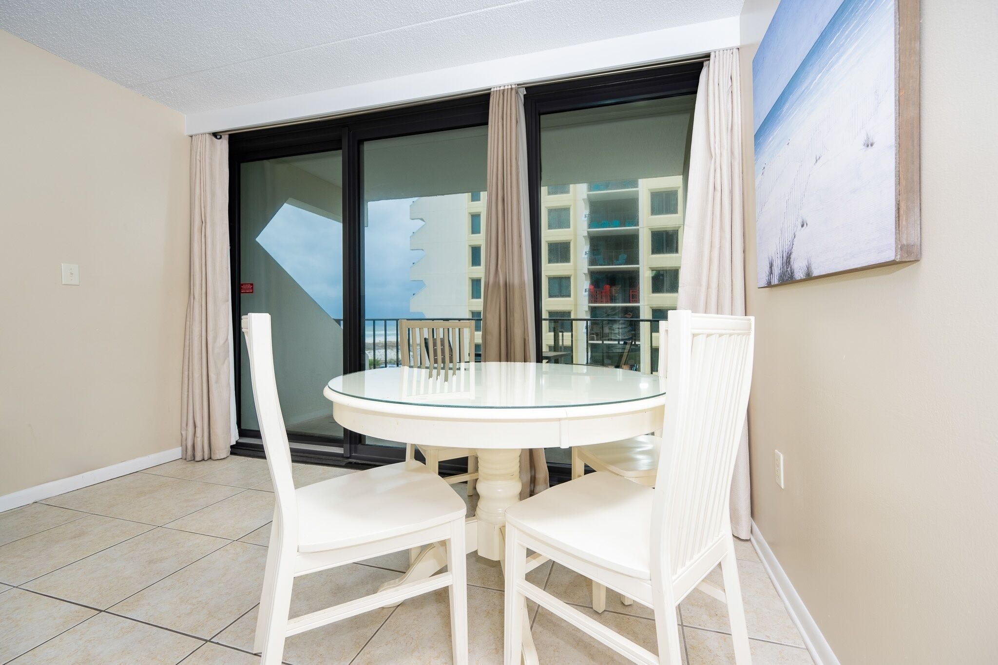 A dining area with a round glass table and white chairs, a sliding glass door leading to a balcony, and a painting on the wall.