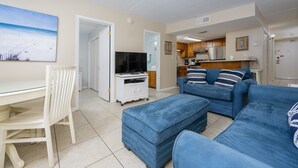 A living room with a TV, a sofa, and a dining area with a chair. - Island Winds East 210 2 Bedroom Condo (Gulf Shores)