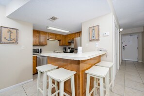 A compact kitchen with wooden cabinets, a central island with stools, a microwave, a refrigerator, and a coffee maker. - Island Winds East 210 2 Bedroom Condo (Gulf Shores)