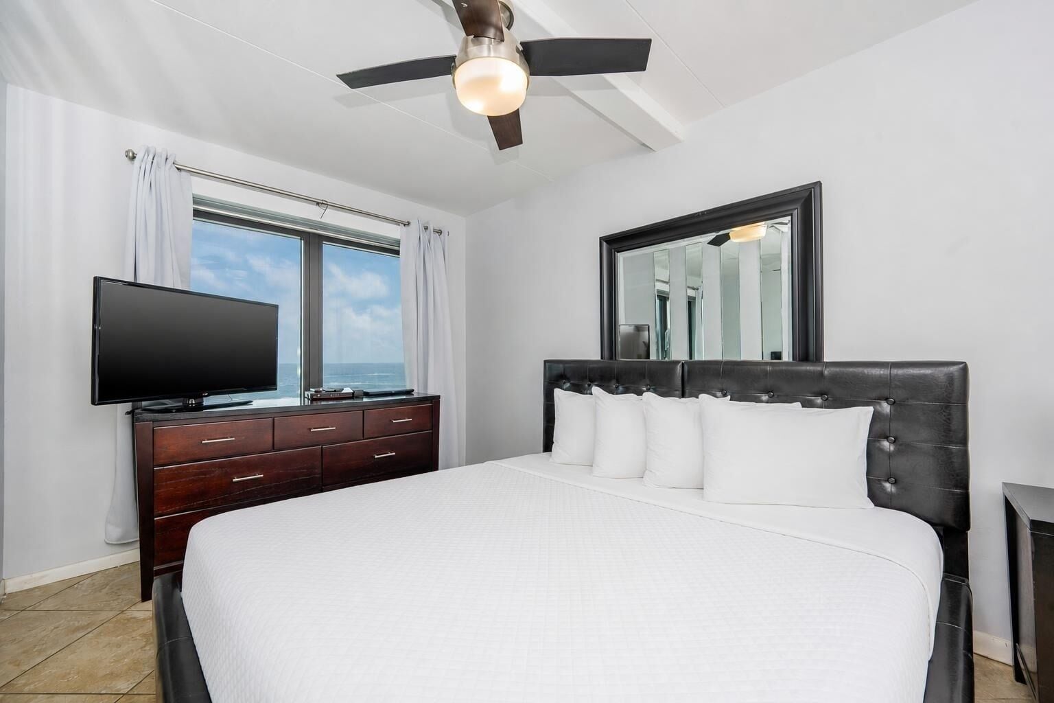 A bedroom with a bed, a dresser, a television, a ceiling fan, and a window with curtains.