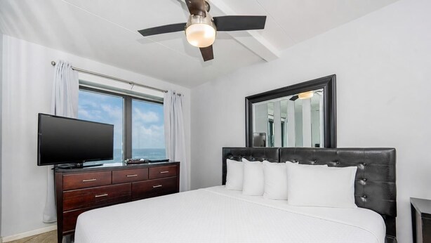 A bedroom with a bed, a dresser, a television, a ceiling fan, and a window with curtains. - Island Winds East 307 2 Bedroom Condo (Gulf Shores)