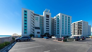 A multi-story hotel with a prominent 'Island Winds East' sign, a parking lot with several cars, and a clear blue sky. - Island Winds East 702 2 Bedroom Condo (Gulf Shores)