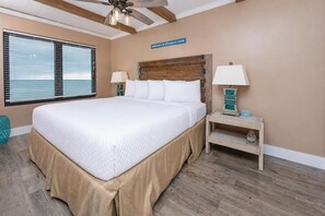 Une chambre avec un grand lit, une tête de lit en bois, une table de chevet avec une lampe et une vue sur l’océan par la fenêtre.