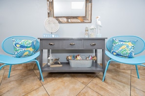 Ein Zimmer mit einer grauen Konsole, zwei blauen Stühlen mit gemusterten Kissen, einem 'WELCOME'-Schild, einem dekorativen Spiegel und einer kleinen Vogelfigur.