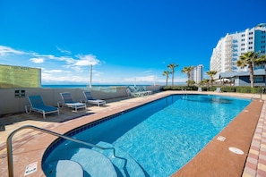 Zona de piscina con sillones de descanso, un cartel con las normas de la piscina y vistas al océano y edificios.