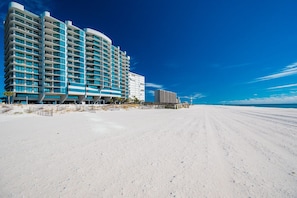 A beachfront hotel with multiple floors, a sandy beach in the foreground, and a clear blue sky. - Phoenix Ix 1002 2 Bedroom Condo (Orange Beach)